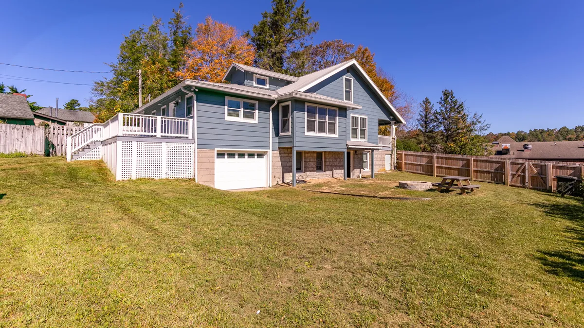 This image shows the backyard with a view of the house’s facade, a spacious deck, the fire pit, and a picnic table set on the lawn. The scene captures a sense of relaxation and communal outdoor living, with a welcoming mood ideal for both day and evening activities.