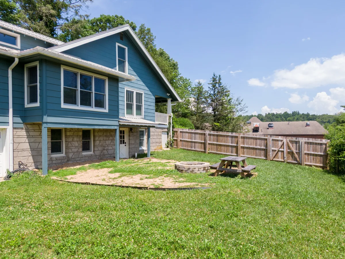 An inviting backyard scene with a lush green lawn, a picnic table for outdoor meals, a fire pit surrounded by chairs, and the blue exterior of the house in the background. The area is clearly fenced, and the cheerful mood suggests a family-friendly space for gatherings and play.