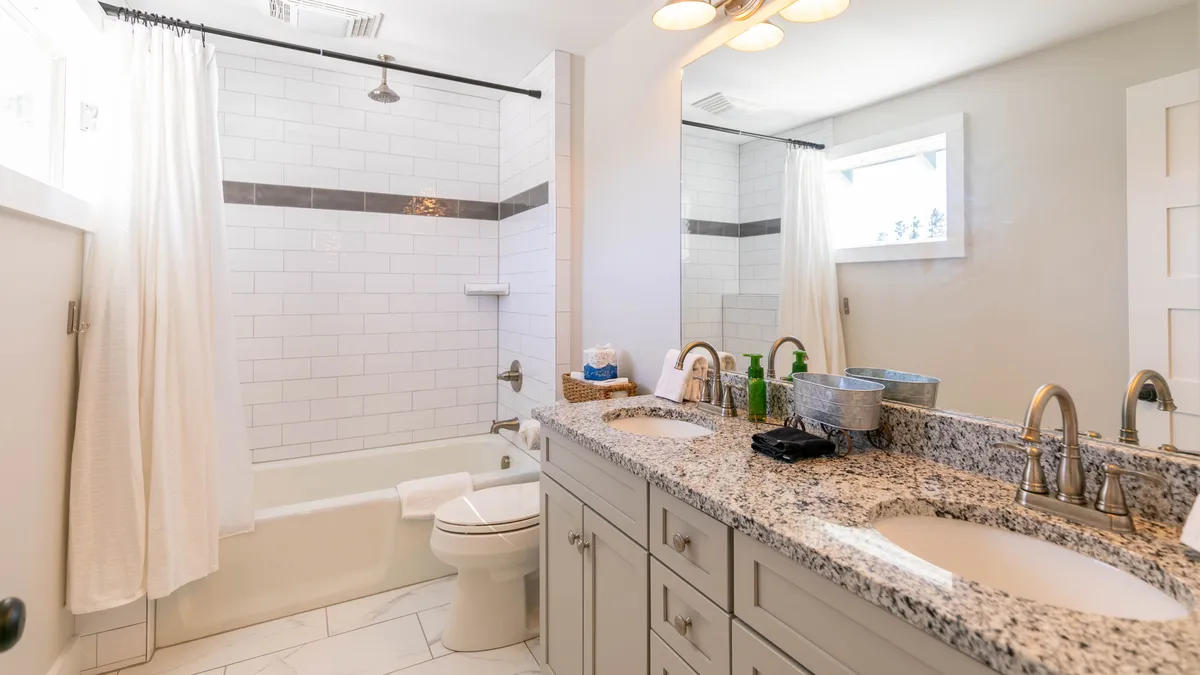 A bright bathroom featuring a granite countertop with double sinks, a bathtub with a shower, and large windows inviting in natural light. The space feels clean, spacious, and perfect for shared use while maintaining a serene ambiance.