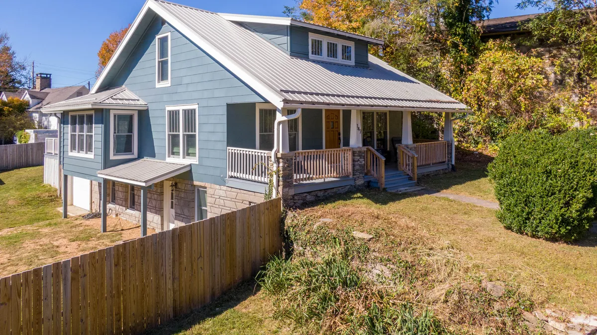 A vibrant view of the blue exterior of the house, highlighting the spacious porch area and verdant landscaping. The scene suggests a space for relaxation and socializing, with the natural setting inviting guests to enjoy the outdoors.
