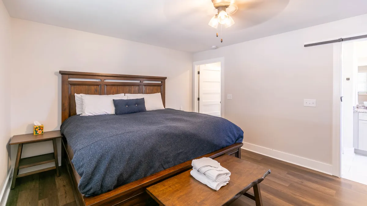 A warmly lit bedroom featuring a wooden bed frame with plush bedding, a nightstand adorned with decorative touches, and a wooden bench at the foot of the bed. The atmosphere is tranquil and inviting, suggesting a restful retreat ideal for winding down at the end of the day.