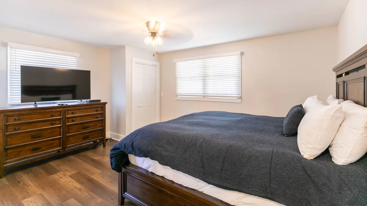 A restful bedroom scene featuring a bed with a dark blanket and crisp white pillows, a wooden dresser, and a television. The mood is calm and inviting, ideal for unwinding, watching TV, or enjoying a peaceful night’s sleep.