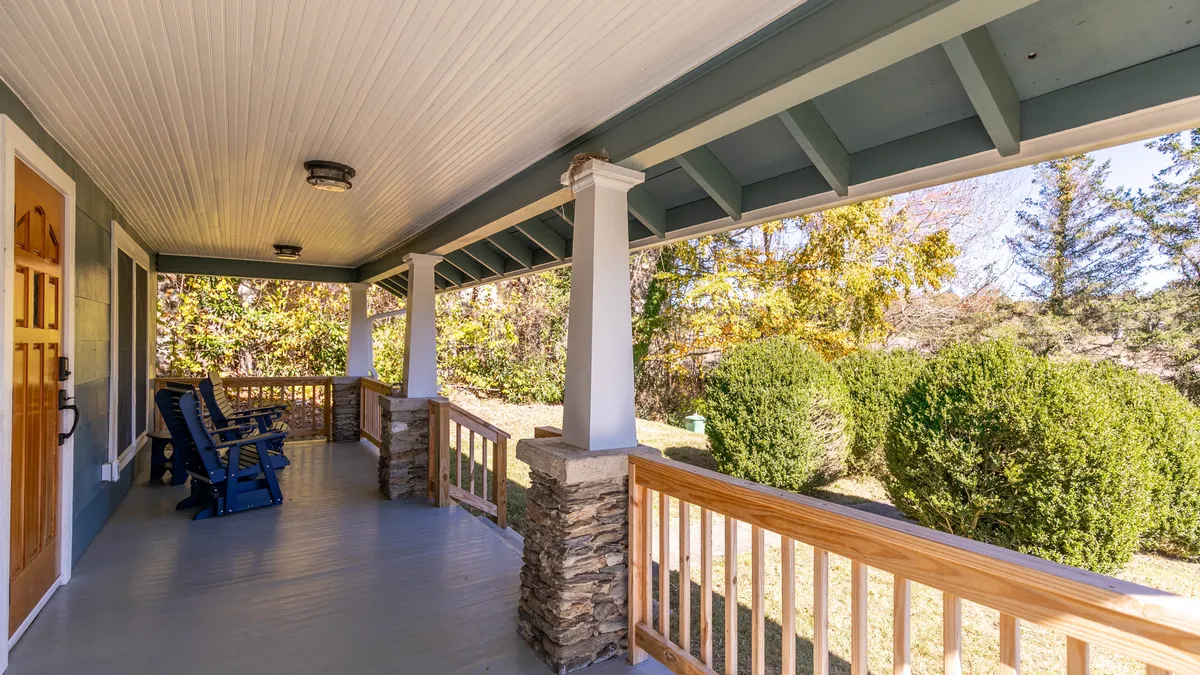 A welcoming front porch with classic rocking chairs lined up against sturdy stone columns, framed by lush greenery. The scene is set at sunset, with soft golden light creating a cozy, inviting atmosphere.